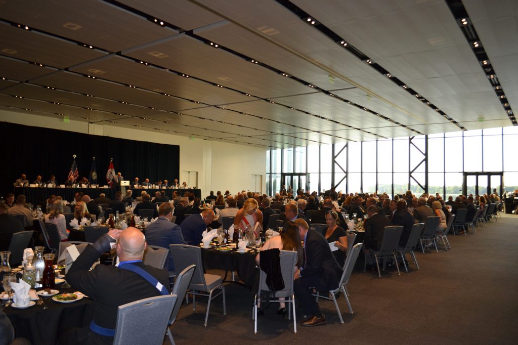 Awards ceremony for Kentucky Association of Police Chiefs conference at the Owensboro Convention Center in Owensboro, Ky. To the left in the background is a raised stage with several people behind a table and someone at the lectern. Many tables filled with people surround several rows back and wide windows of the convention center open to the far right in the distance.